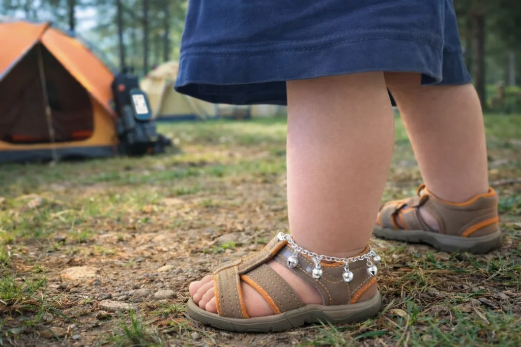 A toddler wearing sturdy footwear and a traditional child anklet (payel/tora) with small bells at a family campsite, illustrating simple, gender-neutral child safety practices during camping in India.